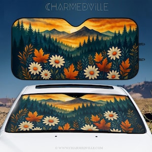 Op de afbeelding: Een auto zonnescherm met een berg- en bosontwerp. Het kunstwerk toont een zonsondergang met oranje en gele tinten, donkergroene bomen, witte madeliefjes en oranje bladeren. De tekst "CHARMEDVILLE" staat bovenaan.