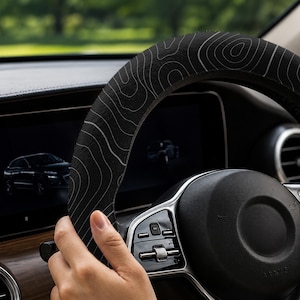 Peut inclure: Housse de volant noire avec un motif de carte topographique blanc. La housse est install&eacute;e sur un volant noir. L'int&eacute;rieur de la voiture est visible en arri&egrave;re-plan.