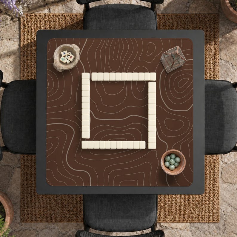 以下が含まれることがあります： ゲーム用にセットされた正方形のテーブルの俯瞰図。テーブルは、地形デザインの濃い茶色の表面を持っています。白いゲームタイルが正方形を形成しています。小さなボウルと幾何学的なガラス容器がテーブルにあります。4つの黒い椅子がテーブルを囲んでいます。