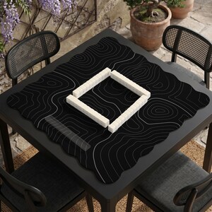 Pu&ograve; includere: Un tappetino da gioco quadrato nero con un motivo a linee topografiche bianche, posizionato su un tavolo scuro. Il tappetino presenta un bordo smerlato e ospita una disposizione quadrata di tessere rettangolari bianche al centro.