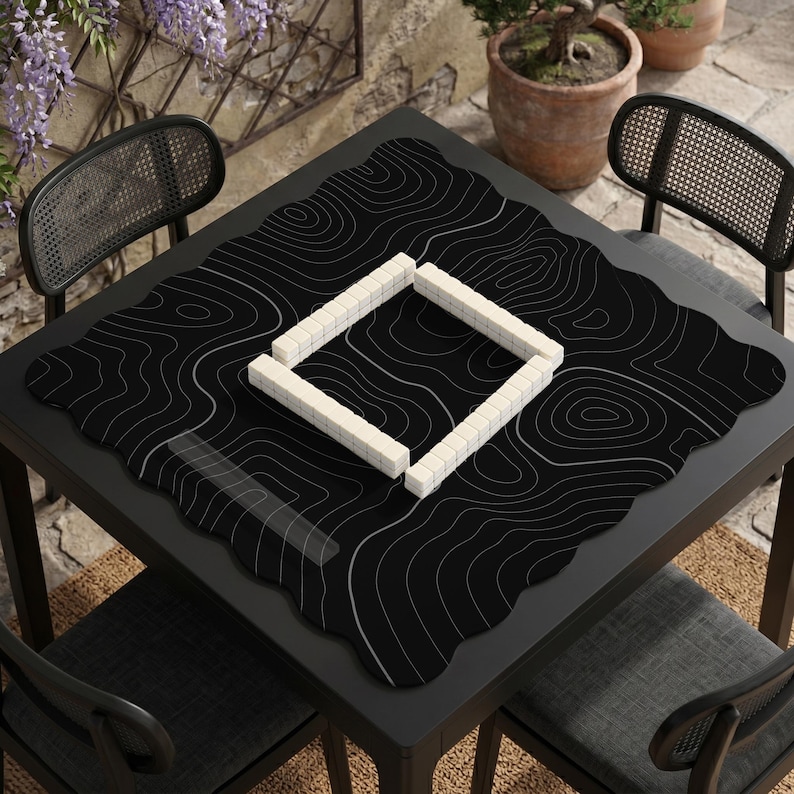 Pu&ograve; includere: Un tappetino da gioco quadrato nero con un motivo a linee topografiche bianche, posizionato su un tavolo scuro. Il tappetino presenta un bordo smerlato e ospita una disposizione quadrata di tessere rettangolari bianche al centro.