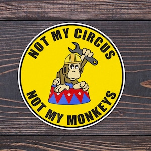 May include: A yellow circular sticker with the text "Not My Circus, Not My Monkeys" in black. A cartoon monkey wearing a hard hat and holding a spanner is sitting on a red, white and blue striped drum.