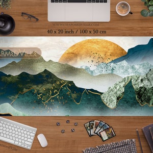 May include: A large, rectangular, white gaming mat with a blue, green, and gold abstract mountain landscape design. The design features a large, gold sun in the upper right corner and a flock of birds flying over the mountains. The mat is 40 inches by 20 inches, or 100 centimeters by 50 centimeters.