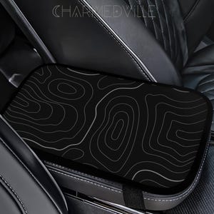 Peut inclure: Housse de console de voiture noire avec un motif de carte topographique blanc. La housse rectangulaire s'adapte sur la console centrale de la voiture. Le texte "CHARMEDVILLE" est visible en haut.