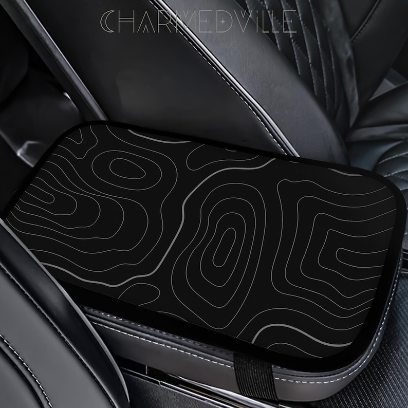 Peut inclure: Housse de console de voiture noire avec un motif de carte topographique blanc. La housse rectangulaire s'adapte sur la console centrale de la voiture. Le texte "CHARMEDVILLE" est visible en haut.