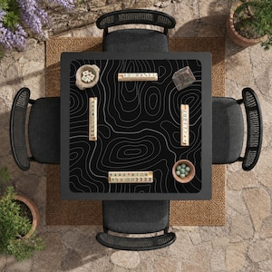 May include: A square black games table featuring a white topographic map pattern on the surface, surrounded by four dark grey chairs. The table holds four wooden tile racks, a small bowl of dice, a bowl of green marbles, and a clear geometric container.