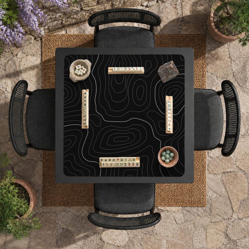Pu&ograve; includere: Un tavolo da gioco quadrato nero con un motivo a mappa topografica bianca sulla superficie, circondato da quattro sedie grigio scuro. Il tavolo contiene quattro supporti per tessere in legno, una ciotola con dadi, una ciotola con biglie verdi e un contenitore geometrico trasparente.