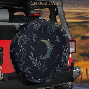 May include: A dark navy blue spare tire cover featuring a crescent moon design with constellations and botanical elements. The cover has a repeating pattern of leaves and stars. The text "CHARMEDVILLE" is visible at the top.