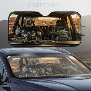 May include: Car sunshade featuring a scene with a Mandalorian character driving a vehicle with Grogu in the passenger seat. The image shows a desert landscape and the text "CHARMEDVILLE" at the top.