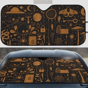 May include: Black and tan car sunshade with a repeating pattern of celestial and mystical elements. The design includes a skull, mushrooms, a moth, a key, a treasure chest, and a dreamcatcher. The text "CHARMEDVILLE" is at the top.