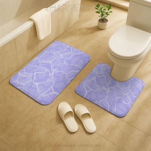 Könnte beinhalten: Ein Badezimmer-Set mit einer rechteckigen Badematte und einer Toilettenkonturmatte, beide mit einem hellvioletten Wasserwellenmuster. Zwei Paar weiße Hausschuhe liegen auf dem Boden. Ein weißes Handtuch hängt am Rand einer Badewanne.