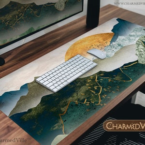 Puede incluir: Un tapete de escritorio con un diseño abstracto de montaña azul, verde y dorado. El tapete presenta un patrón de sol dorado y blanco en la esquina superior derecha. Un teclado y un ratón inalámbricos blancos están sobre el tapete. El texto "CHARMEDVILLE" está en la esquina inferior derecha.