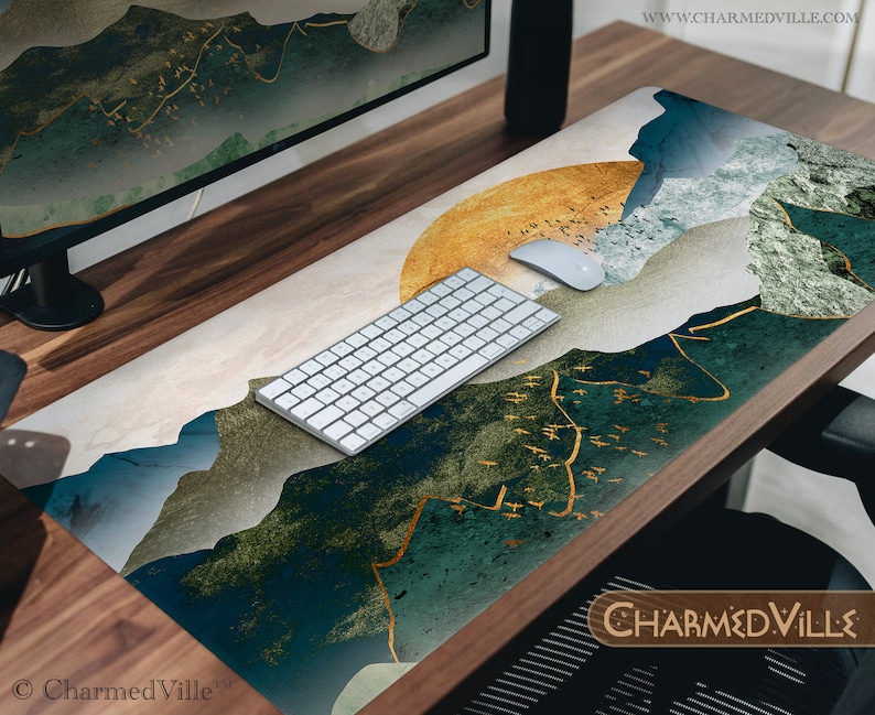 May include: A desk mat with a blue, green, and gold abstract mountain design. The mat features a gold and white sunburst pattern in the upper right corner. A white wireless keyboard and mouse are on the mat. The text "CHARMEDVILLE" is in the lower right corner.