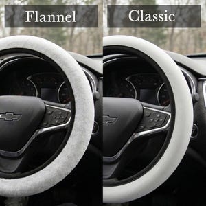 Peut inclure: Deux housses de volant, l'une avec une texture Flannel blanche et duveteuse, l'autre avec une texture Classic blanche et lisse. Les housses sont pr&eacute;sent&eacute;es sur un volant noir avec des accents argent&eacute;s. Le tableau de bord est visible.
