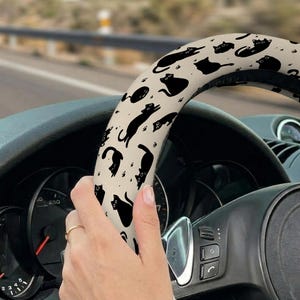 May include: A beige steering wheel cover featuring a repeating pattern of black cat silhouettes and paw prints. The cover is wrapped around a black steering wheel, with a hand holding the wheel. The background shows a road and a car dashboard.