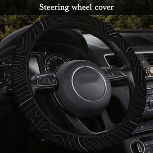 Peut inclure: Housse de volant noire avec un motif topographique blanc. La housse est install&eacute;e sur le volant d'une voiture. Le texte "Steering wheel cover" est visible en haut de l'image.