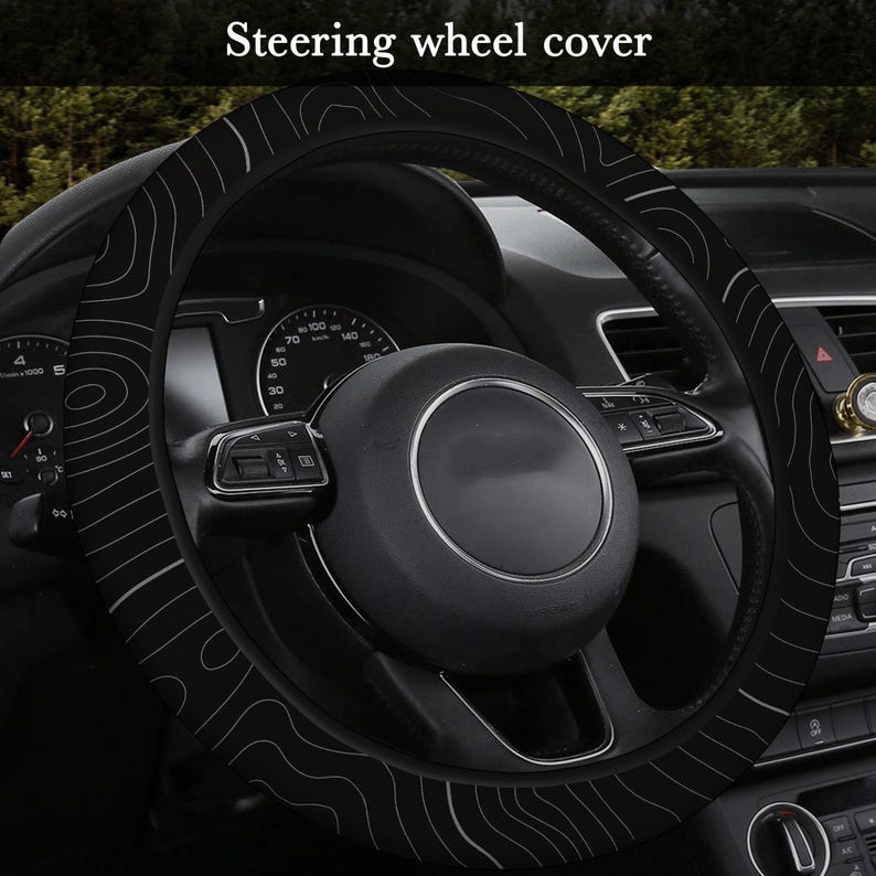 Peut inclure: Housse de volant noire avec un motif topographique blanc. La housse est install&eacute;e sur le volant d'une voiture. Le texte "Steering wheel cover" est visible en haut de l'image.