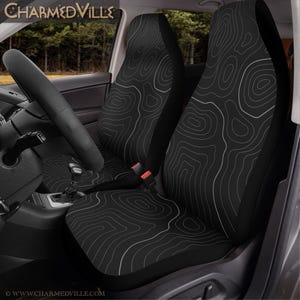 Peut inclure: Housses de si&egrave;ge de voiture noires avec un motif de carte topographique blanc. L'image montre les housses install&eacute;es dans une voiture, avec le volant et le tableau de bord visibles. Le texte "CHARMEDVILLE" est en haut.