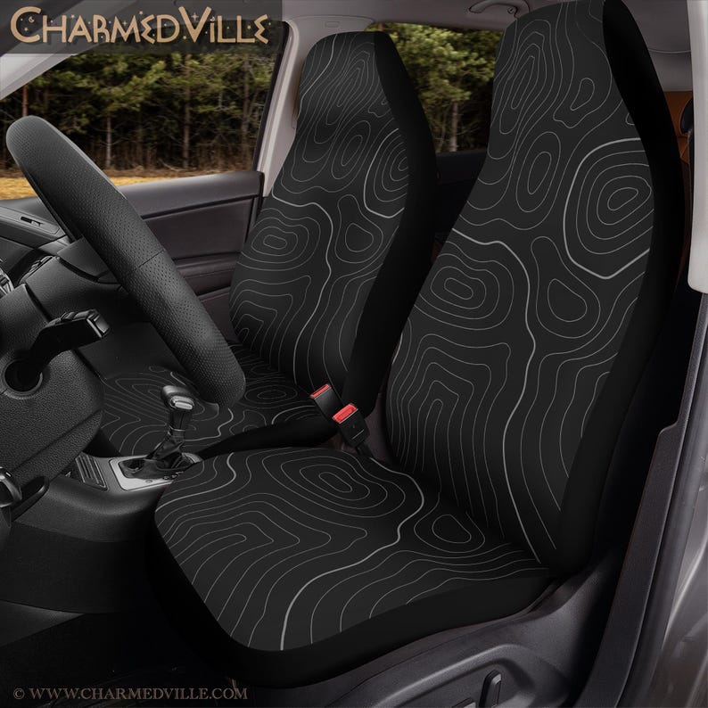 Peut inclure: Housses de si&egrave;ge de voiture noires avec un motif de carte topographique blanc. L'image montre les housses install&eacute;es dans une voiture, avec le volant et le tableau de bord visibles. Le texte "CHARMEDVILLE" est en haut.