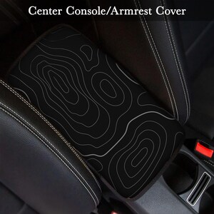 Peut inclure: Housse de console centrale/accoudoir noire avec un motif de carte topographique blanc. La housse est rectangulaire et s'adapte sur l'accoudoir d'une voiture. Le texte "Center Console/Armrest Cover" est en haut de l'image.