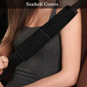 Peut inclure: Housse de ceinture de s&eacute;curit&eacute; noire avec un motif topographique blanc. Le texte "Seatbelt Covers" est visible en haut de l'image. La housse est fix&eacute;e &agrave; une ceinture de s&eacute;curit&eacute; noire. Une personne porte la ceinture.