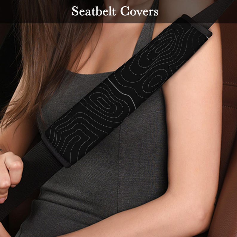 Peut inclure: Housse de ceinture de s&eacute;curit&eacute; noire avec un motif topographique blanc. Le texte "Seatbelt Covers" est visible en haut de l'image. La housse est fix&eacute;e &agrave; une ceinture de s&eacute;curit&eacute; noire. Une personne porte la ceinture.