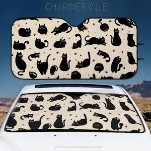 Op de afbeelding: Auto zonnescherm met een herhalend patroon van zwarte kat silhouetten op een crème achtergrond, met pootafdrukken. De tekst "CHARMEDVILLE" staat bovenaan. Het zonnescherm is geïnstalleerd in een witte auto.