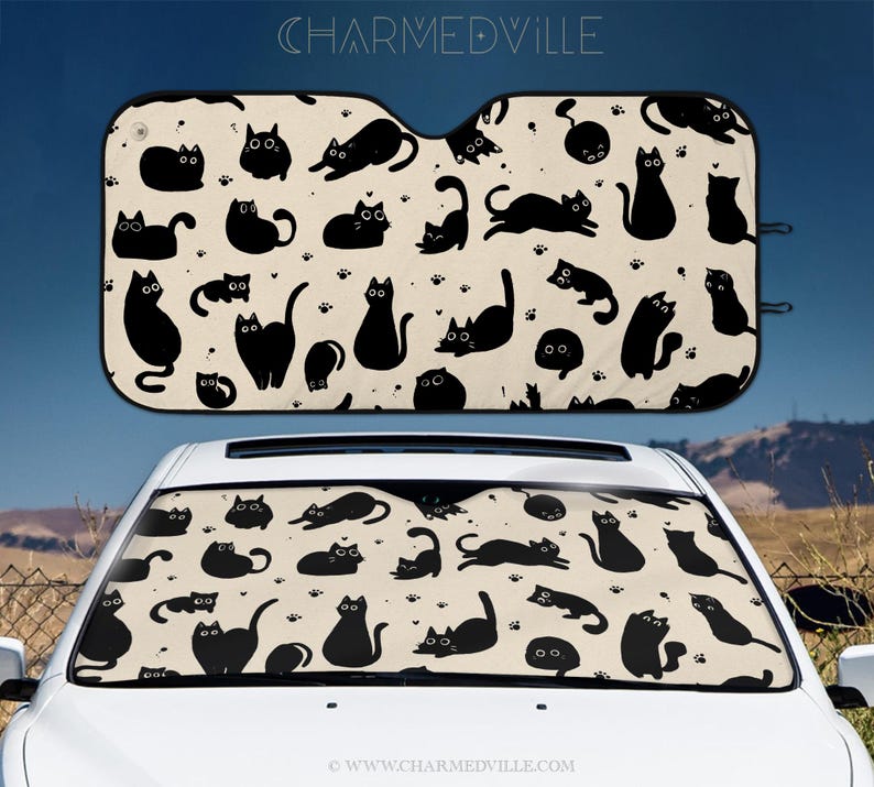 May include: Car sunshade with a repeating pattern of black cat silhouettes on a cream background, with paw prints. The text "CHARMEDVILLE" is at the top. The sunshade is installed in a white car.
