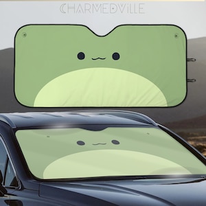 May include: A green car sunshade with a cartoon frog design. The frog has a light green body and a dark green face with black eyes and a small smile.