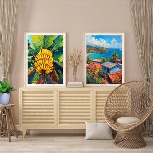 May include: Two framed art prints. One shows a bunch of yellow bananas with green leaves. The other depicts a coastal scene with blue water, red-roofed buildings, and palm trees. The artwork is displayed on a light wood cabinet.
