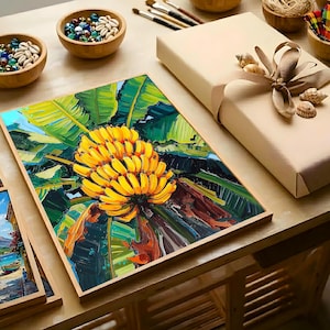 May include: A framed painting of a bunch of yellow bananas with green leaves. The artwork is displayed on a wooden table, alongside a wrapped gift box, bowls of shells, and art supplies. The painting has a vibrant, textured appearance.