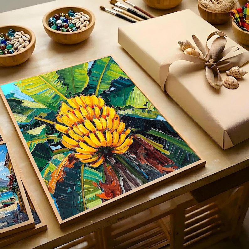 May include: A framed painting of a bunch of yellow bananas with green leaves. The artwork is displayed on a wooden table, alongside a wrapped gift box, bowls of shells, and art supplies. The painting has a vibrant, textured appearance.