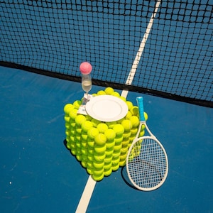 May include: A tennis-themed display on a blue court. A cube made of tennis balls supports a white plate, a champagne glass with a pink garnish, and a tennis racket. The net is in the background.