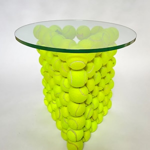 Puede incluir: Una mesa única y llamativa con una tapa de vidrio transparente sostenida por una base hecha completamente de pelotas de tenis de color amarillo brillante. Las pelotas de tenis están dispuestas en una forma triangular apilada, creando una estética lúdica y deportiva. La mesa mide unos 75 cm de alto.