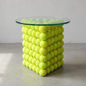 May include: A side table featuring a square base constructed from numerous bright yellow tennis balls stacked in a grid pattern, topped with a circular, clear glass tabletop that provides a flat surface for placing various household items.