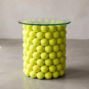 May include: A cylindrical side table constructed from numerous bright yellow tennis balls stacked in a vertical formation, topped with a circular, clear glass surface that provides a flat, stable area for placing items on the structure.