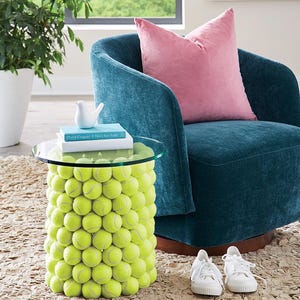 May include: A unique side table made from tennis balls, topped with a clear glass surface. A teal armchair with a pink pillow sits nearby. White sneakers are on the floor. A small white bird figurine and books are on the table.