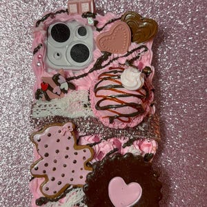 May include: A phone case decorated with pink whipped cream texture, featuring a pink teddy bear cookie, a dark chocolate biscuit with a pink heart center, a pink ice cream scoop, heart-shaped charms, and white lace trim accents.