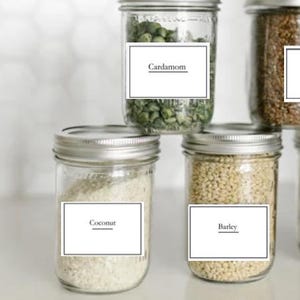 May include: Four glass jars with white labels and black text. The labels read "Cardamom", "Coconut", "Barley", and an unreadable label. The jars are filled with various grains and seeds.