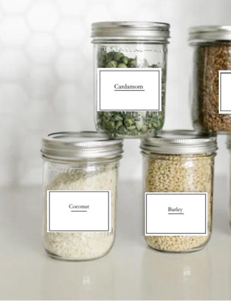 May include: Four glass jars with white labels and black text. The labels read "Cardamom", "Coconut", "Barley", and an unreadable label. The jars are filled with various grains and seeds.