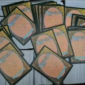 May include: A box filled with Magic: The Gathering trading cards. The cards are all the same, with a brown and gold border and a multi-colored symbol in the center. The box is made of cardboard and has a brown color.