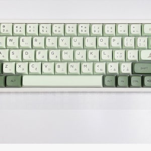 Könnte beinhalten: Eine weiße und grüne mechanische Tastatur mit japanischen Tasten. Die Tastatur hat eine hellgrüne Leertaste und eine dunkelgrüne Escape-Taste. Die Tastatur ist in der oberen rechten Ecke mit "Matcha" beschriftet.
