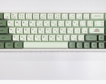 以下が含まれることがあります： 日本語のキーキャップが付いた白と緑のメカニカルキーボード。キーボードは、薄い緑色のスペースバーと濃い緑色のエスケープキーを持っています。キーボードは右上の隅に「抹茶」と表示されています。