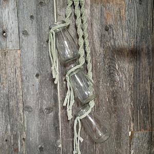 May include: A hanging planter featuring three clear glass jars suspended by a light green macrame rope. The macrame is attached to a wooden ring at the top. The jars are tied with matching rope.