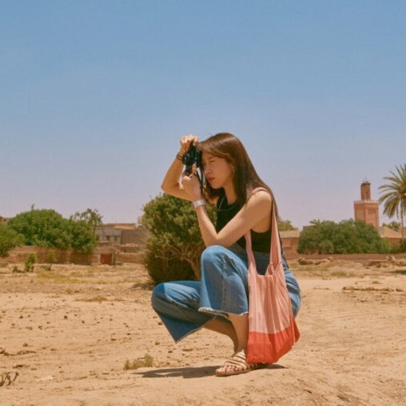 May include: A woman wearing blue jeans and a black tank top is squatting and taking a photo with a camera. She is wearing a pink and red tote bag.
