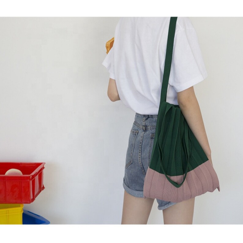 May include: A green and pink knitted tote bag with a long strap. The bag is pleated and has a casual, relaxed style.