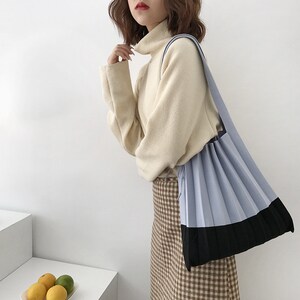 May include: A light blue and black pleated tote bag with a long strap. The bag is being worn by a person wearing a cream turtleneck sweater and a brown and white checked skirt.