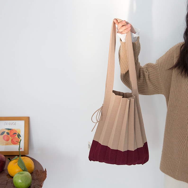 May include: A beige and burgundy pleated tote bag with a long strap. The bag is made of a soft, textured fabric and has a simple, elegant design.