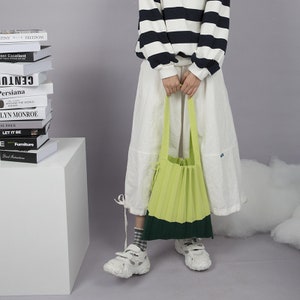 May include: A green and dark green pleated tote bag with a long strap. The bag is being held by a person wearing a white skirt and a black and white striped sweater.