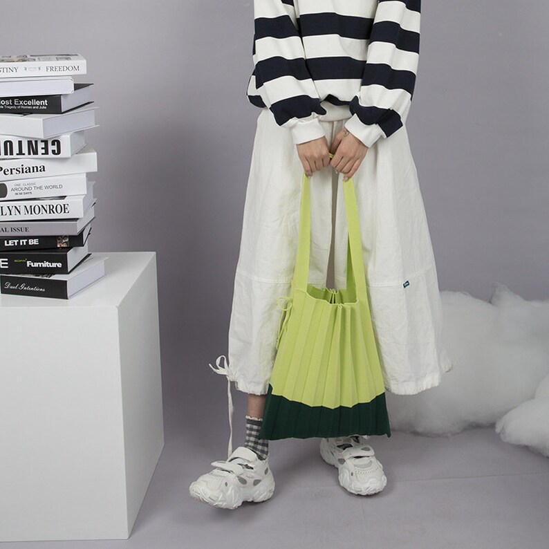 May include: A green and dark green pleated tote bag with a long strap. The bag is being held by a person wearing a white skirt and a black and white striped sweater.